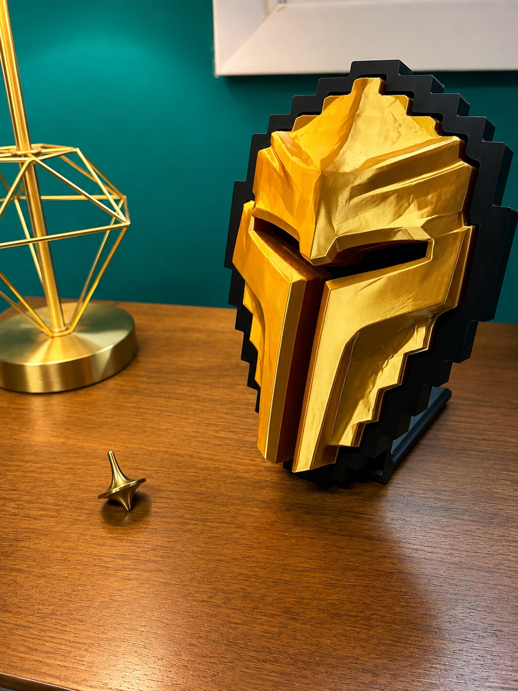 A Tibia Golden Helmet display stand on a wooden table next to a gold geometric lamp, set against a teal wall.