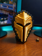 A 3D-printed Tibia Golden Helmet in a pixelated block design, finished in metallic gold with a black pixelated border, displayed on its stand on top of a wooden desk.